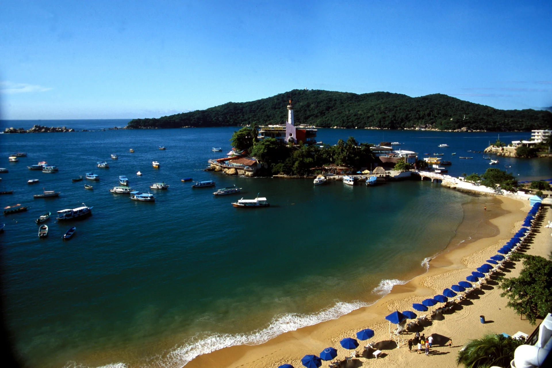 La playa Caleta en curva, con lanchas en la ensenada, el islote de Mágico Mundo Marino y la isla La Roqueta al fondo, Playas Caleta y Caletilla, Guerrero