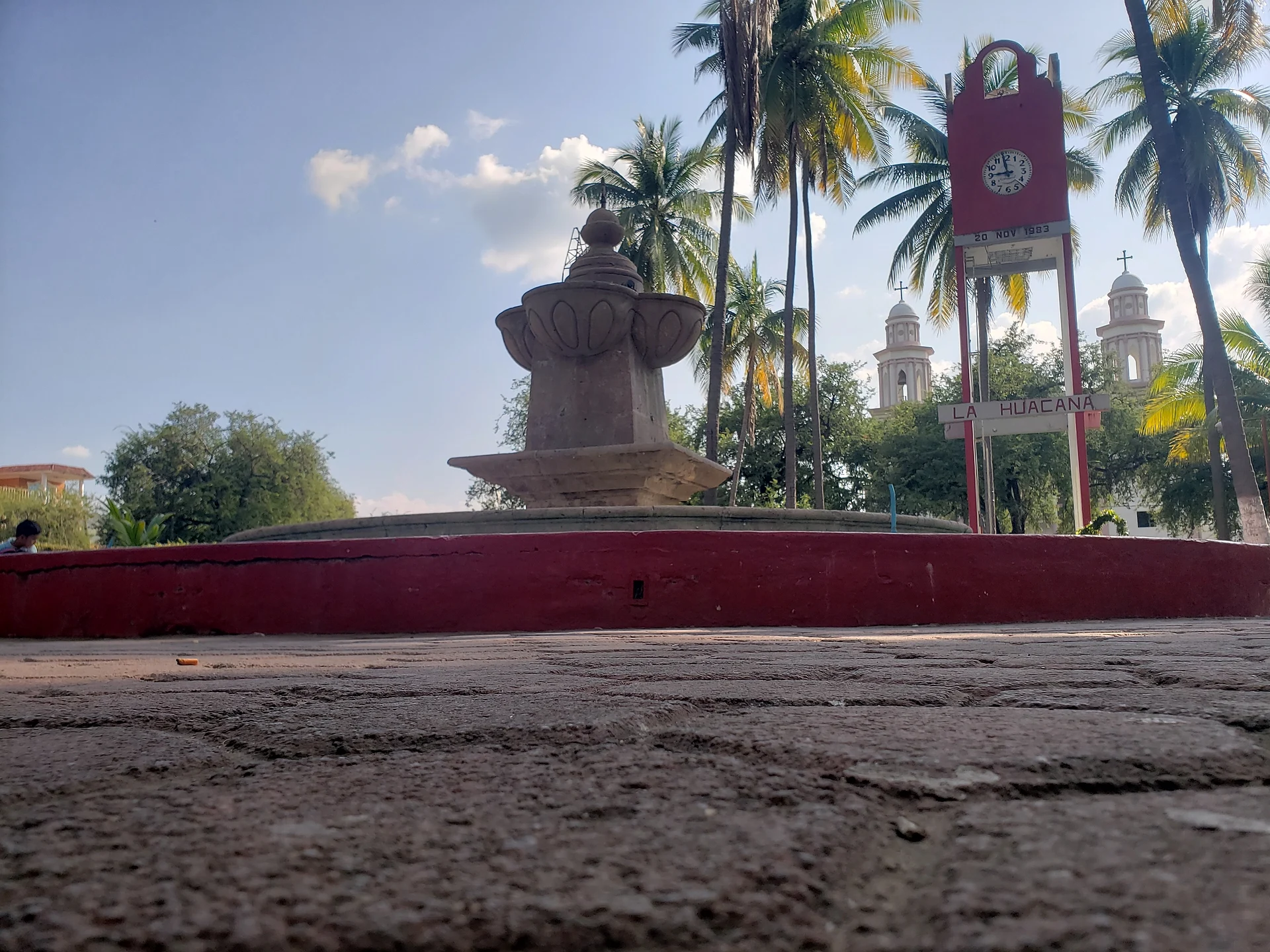 Fuente de cantera en la plaza principal de La Huacana, con palmeras, la torre roja del reloj y las torres de la parroquia al fondo, Michoacán