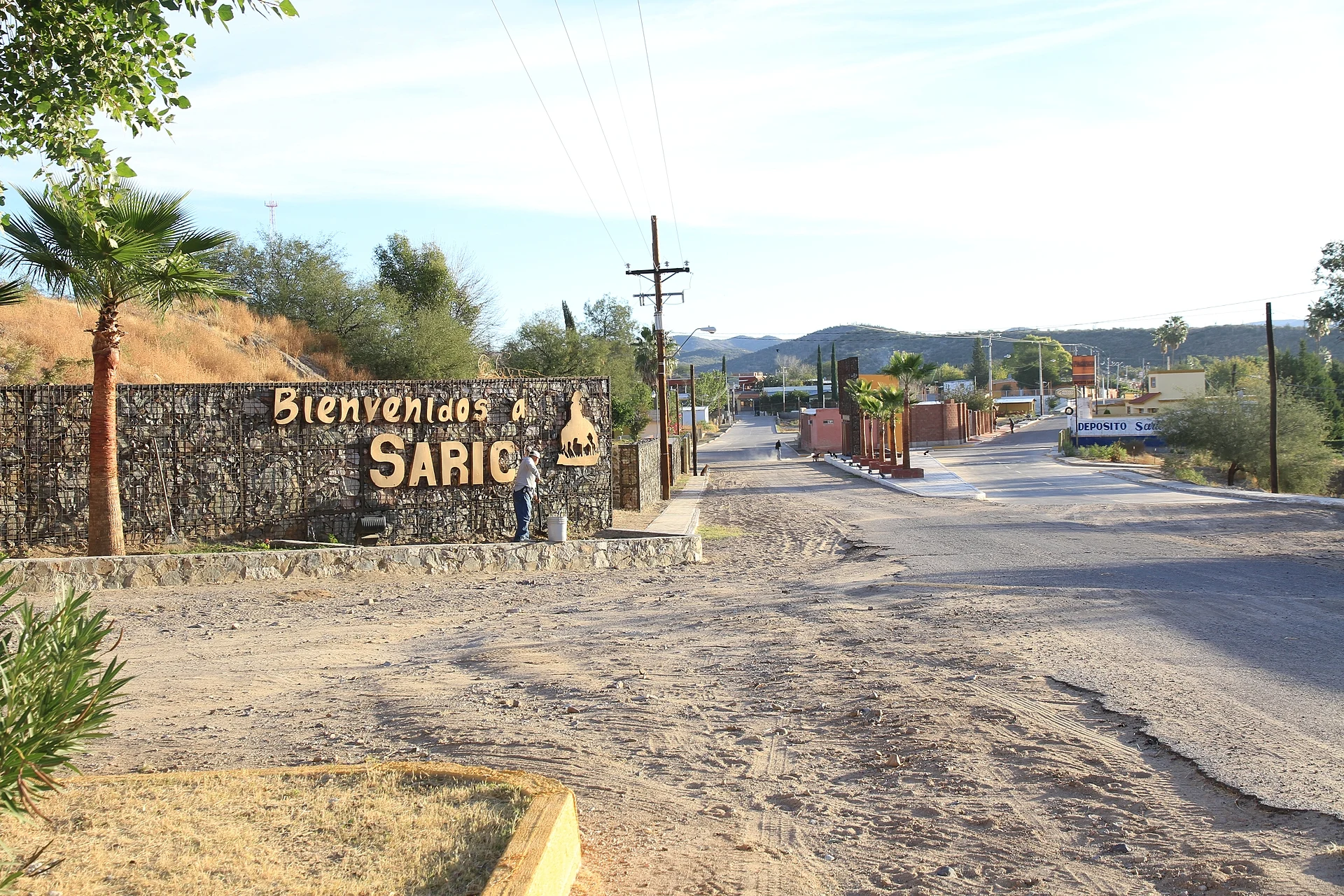 El muro de piedra con el letrero "Bienvenidos a Sáric" a la entrada del pueblo, con palmas, la calle pavimentada y cerros del desierto al fondo, Sonora