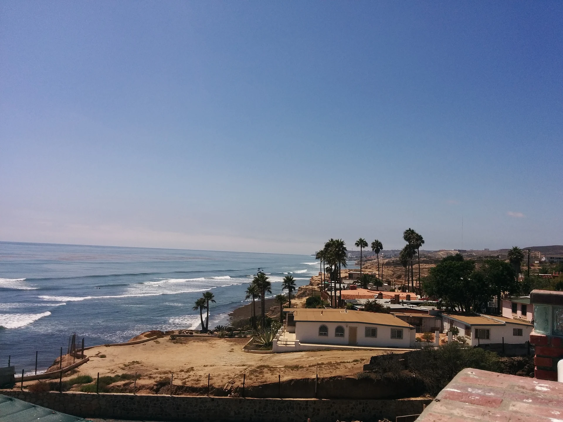 El acantilado bajo de Puerto Nuevo sobre el Pacífico visto desde una azotea, con palmeras, casas blancas y olas rompiendo en la orilla, Baja California