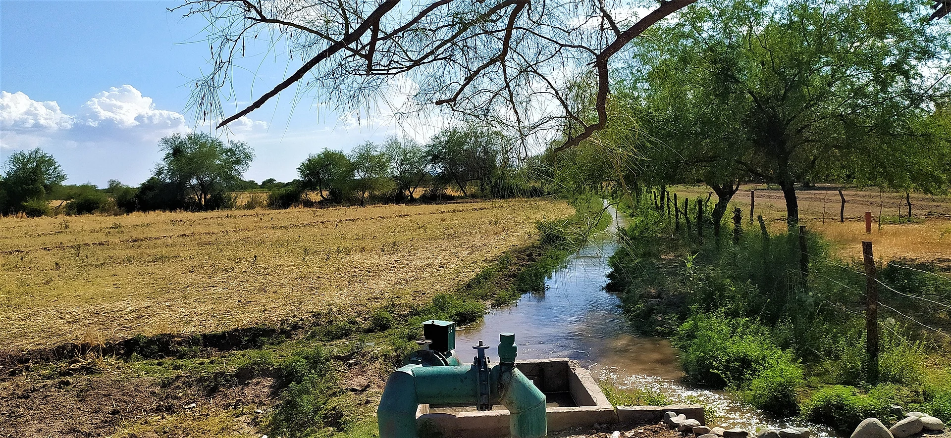 Un canal de riego del ejido Molino de Camou junto a una toma de bomba verde, entre una parcela seca ya cosechada y mezquites tras un cerco de alambre, Sonora
