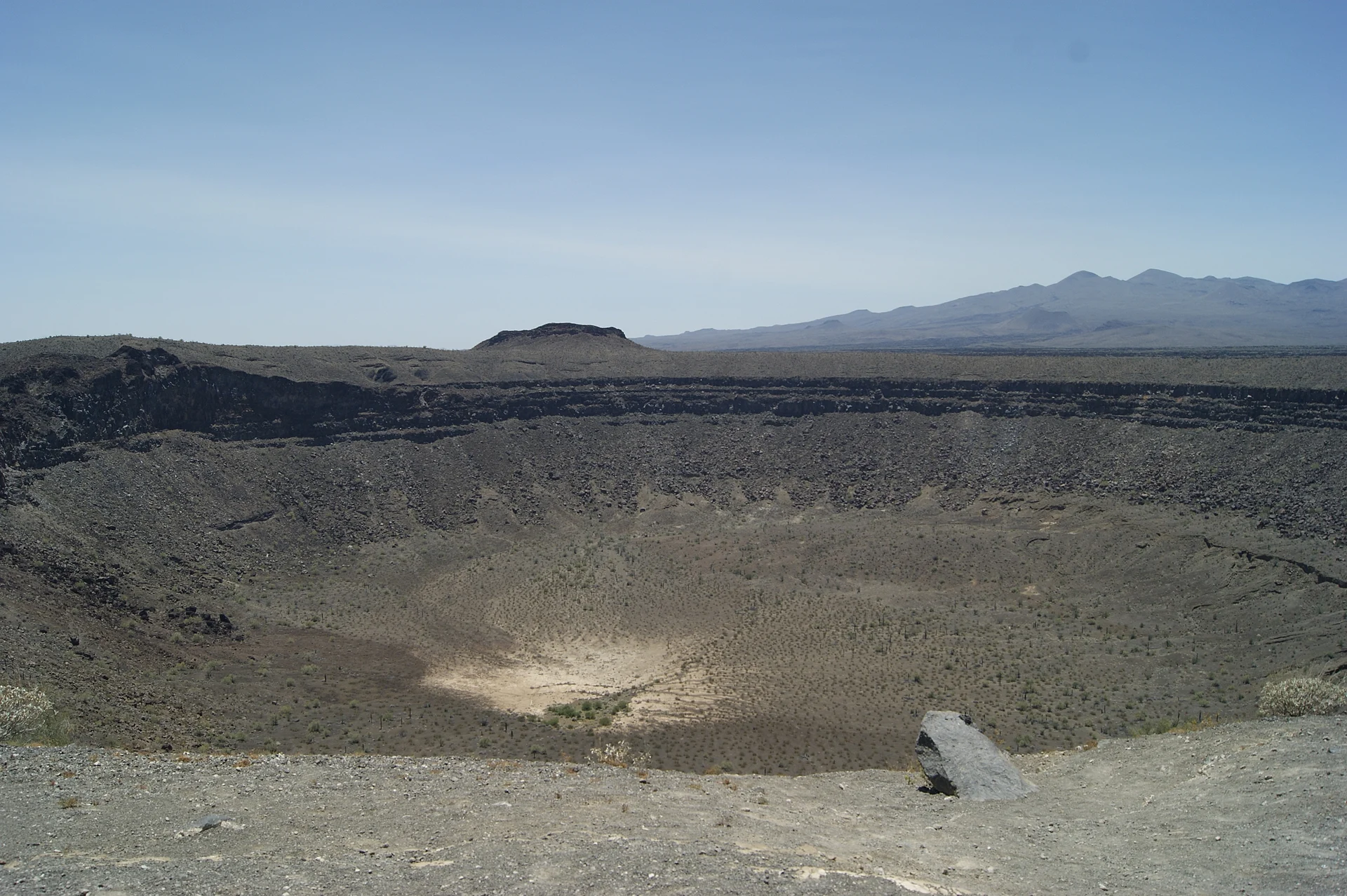 Un enorme cráter volcánico redondo de El Pinacate visto desde el borde, con paredes de capas de lava oscura que bajan a un fondo de arena clara y sierras del desierto al fondo, Reserva de la Biosfera El Pinacate y Gran Desierto de Altar, Sonora