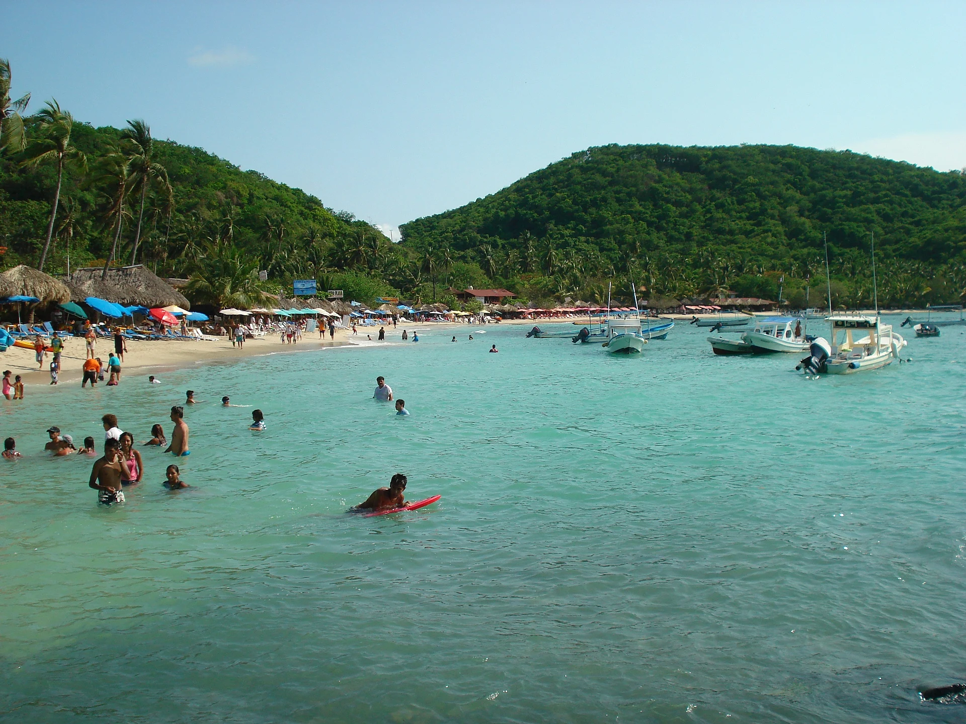 Bañistas en el agua turquesa y tranquila de Playa Las Gatas, con palapas en la arena y cerros verdes al fondo, Guerrero