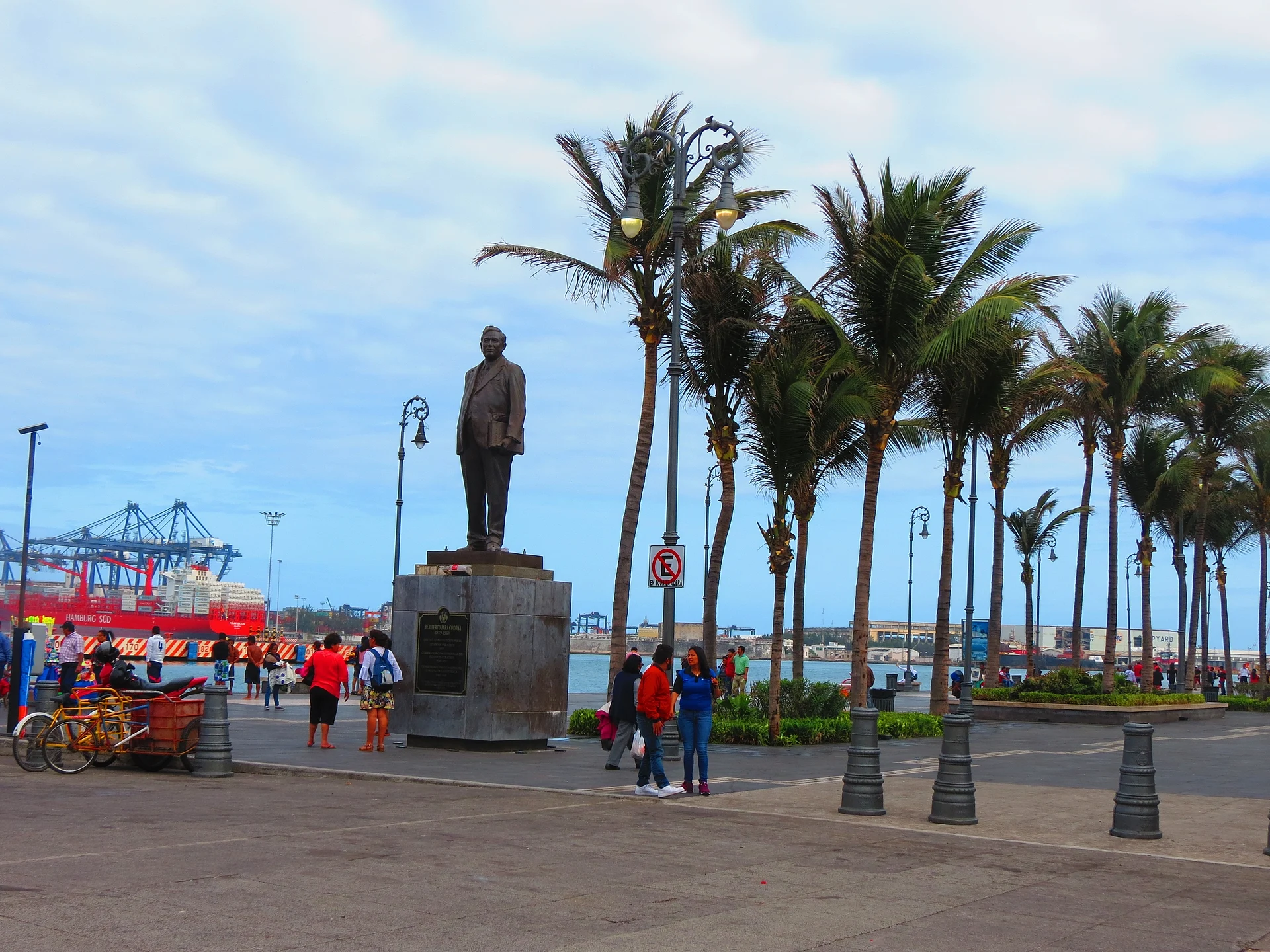 Palm-lined Malecón de Veracruz with a bronze statue, strollers and port cranes in the background
